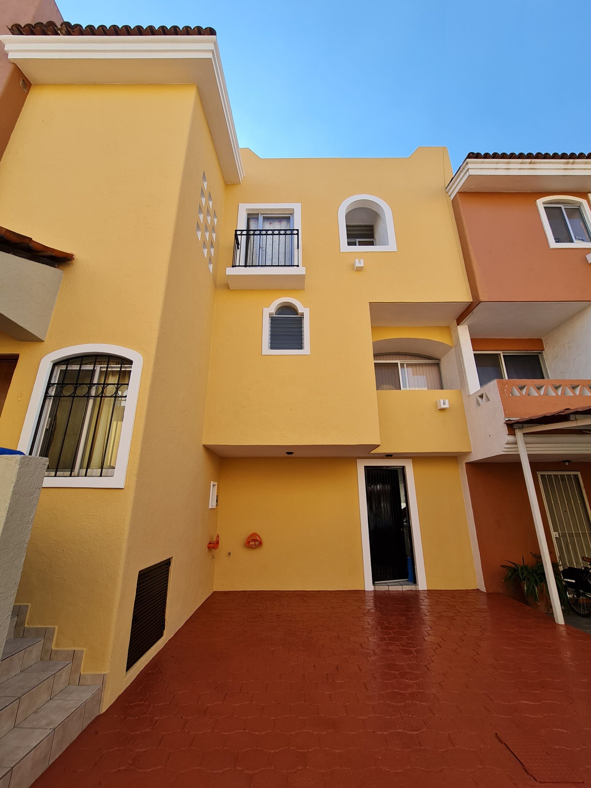 Una casa amarilla de tres pisos con estilo moderno y entradas arqueadas, paredes lisas, ubicada en un vecindario soleado, sin elementos históricos visibles.