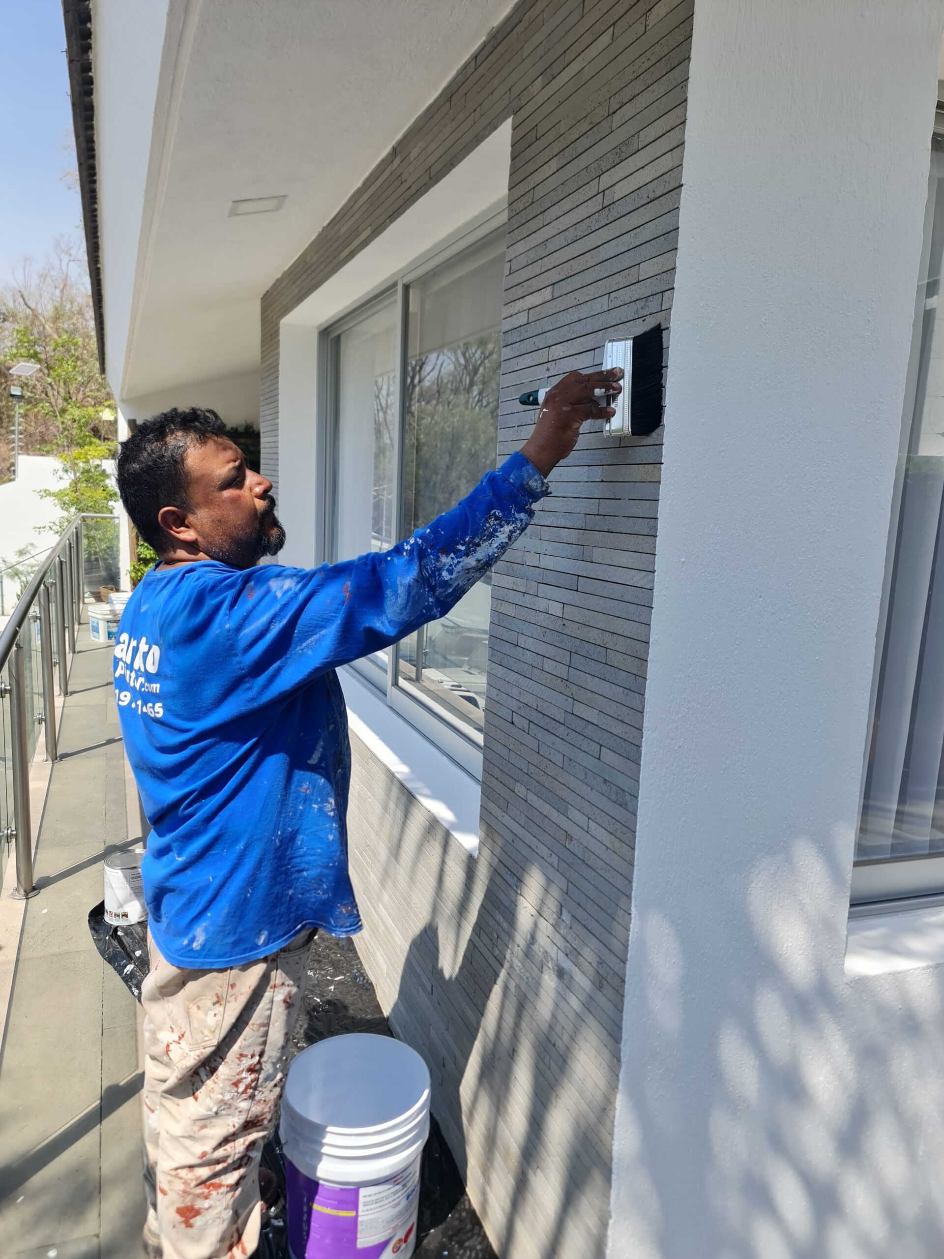 Una persona pinta una pared exterior con esponja, vestido en azul. Hay cubetas de pintura y ventanas grandes al fondo.